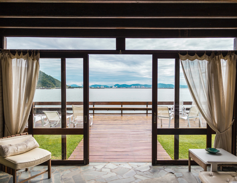 View looking out of a room that is on the water with floor to ceiling glass and wooden doors that has mountains and a city in the background