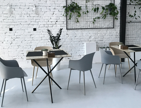 Grey and tan chairs and tables in restaurant setting with a brick wall painted white that has hanging plants and light grey flooring