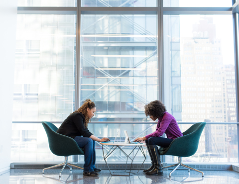 Two women sitting at a table on their laptops in a lobby, overlooking a city street